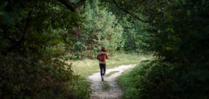 Anonymous distant backpacker with rucksack strolling on trail in woods with lush green trees during trekking in nature on summer day