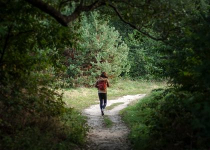 Anonymous distant backpacker with rucksack strolling on trail in woods with lush green trees during trekking in nature on summer day