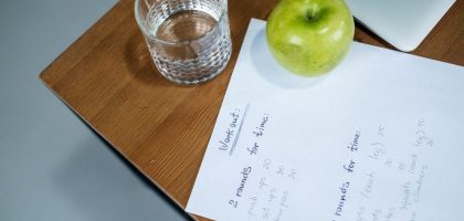 A green apple, glass of water, and workout plan on a wooden desk symbolize healthy living.
